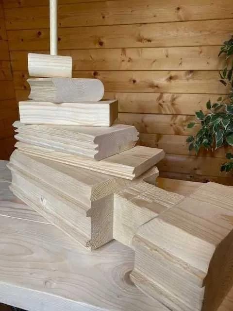 Elementy konstrukcyjne z jasnego drewna ułożone warstwowo, prawdopodobnie do budowy domu z bali lub sauny, widoczne detale połączeń i obróbki drewna.