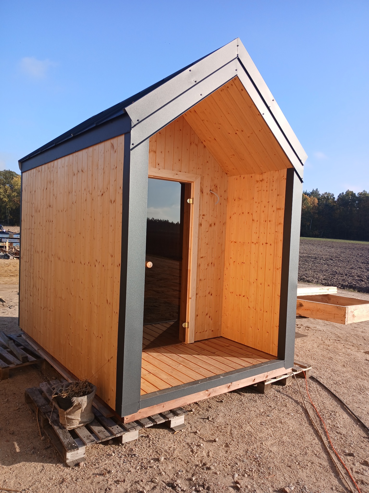 Prosty, drewniany domek z ciemną ramą i szklanymi drzwiami, umieszczony na paletach na piaszczystym terenie, z widocznym polem i drzewami w tle.