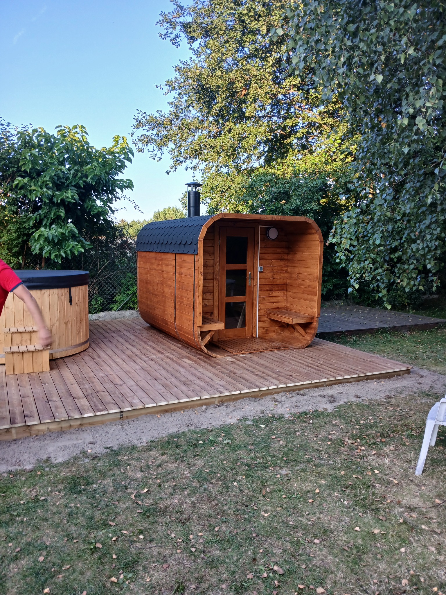 Drewniana beczka z hydromasażem i beczkowa sauna z kominem na tarasie z desek, otoczone zielenią.