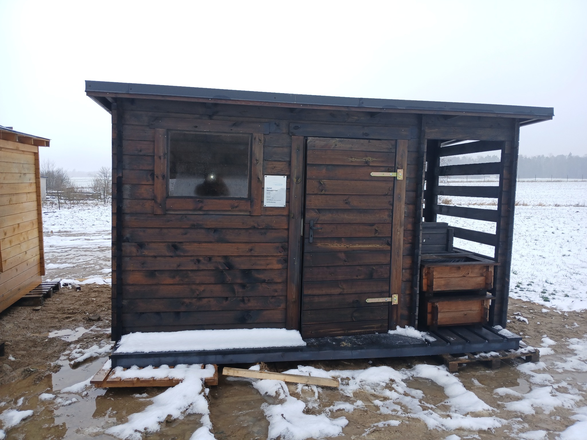 Ciemna, drewniana sauna zewnętrzna z oknem i otwartą przestrzenią na drewno, ustawiona na paletach na zasypanym śniegiem terenie.