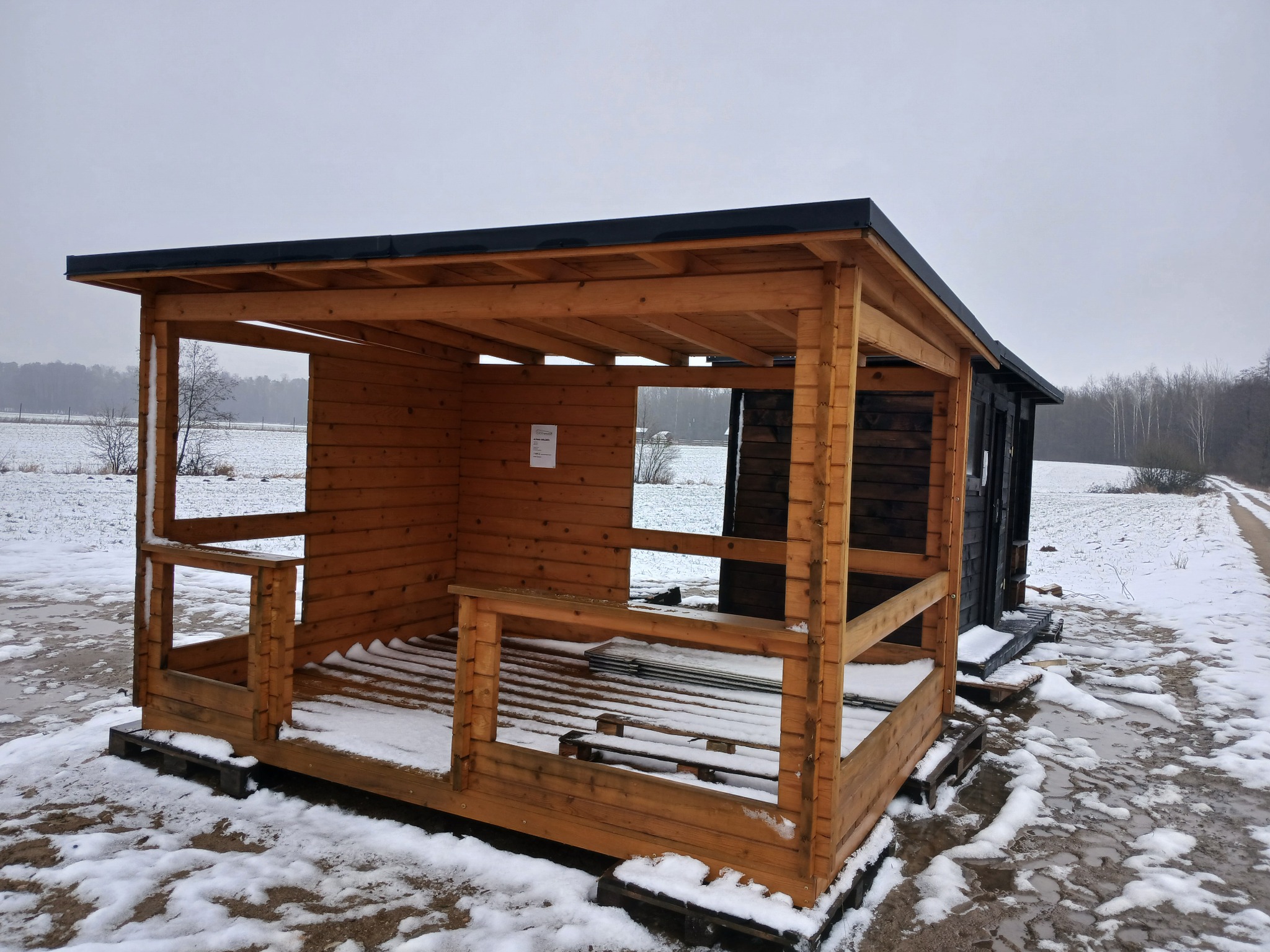 Surowa drewniana konstrukcja z czarnym dachem na paletach, częściowo obita deskami, stojąca na śniegu na tle zaśnieżonego pola i lasu.
