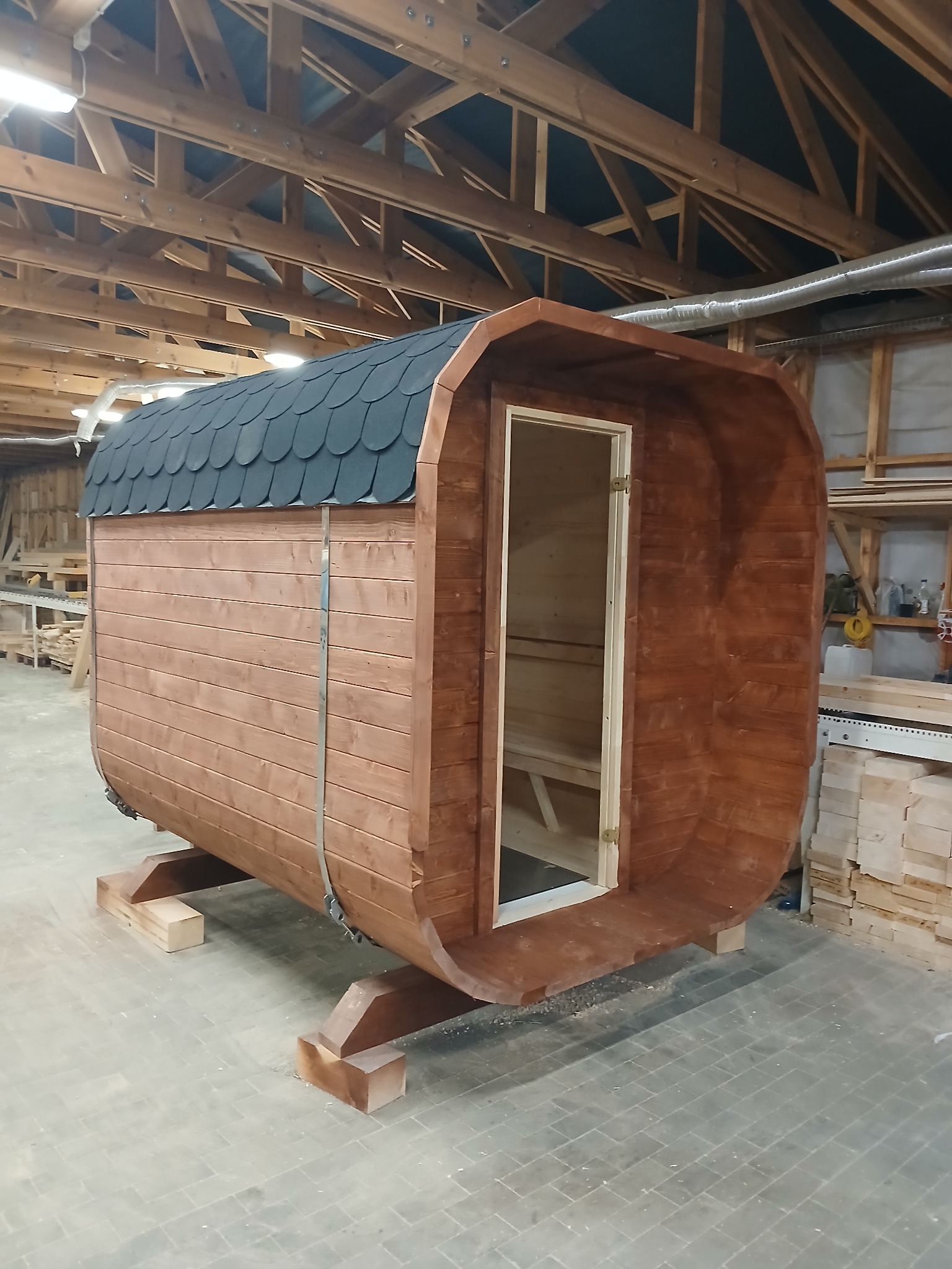 Gotowa beczka sauny z drewnianą konstrukcją i czarnym dachem z gontu bitumicznego, umieszczona w hali produkcyjnej na drewnianych podporach.