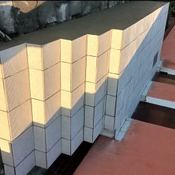 Osoba prywatna - Wznoszenie ściany z bloczków betonowych o ryflowanej powierzchni, widoczne połączenia zaprawą, fragment budynku z oknami w tle, oświetlone popołudniowym słońcem.