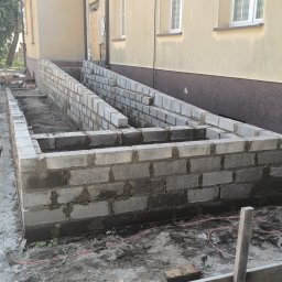 Osoba prywatna - Wznoszenie ścian fundamentowych z bloczków betonowych przy budynku, widoczne zaprawy i roślinność wokół