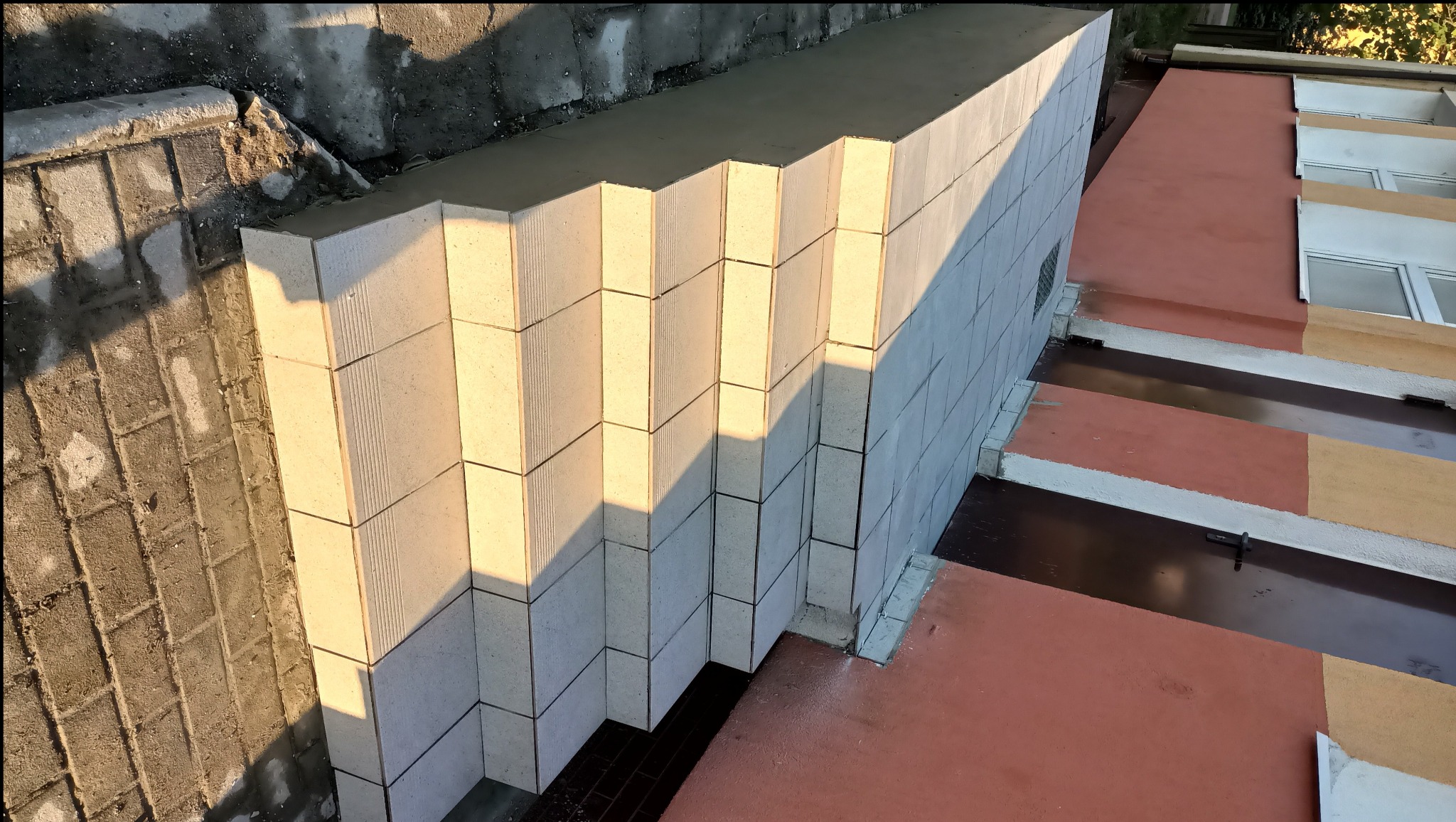 Wznoszenie ściany z bloczków betonowych o ryflowanej powierzchni, widoczne połączenia zaprawą, fragment budynku z oknami w tle, oświetlone popołudniowym słońcem.