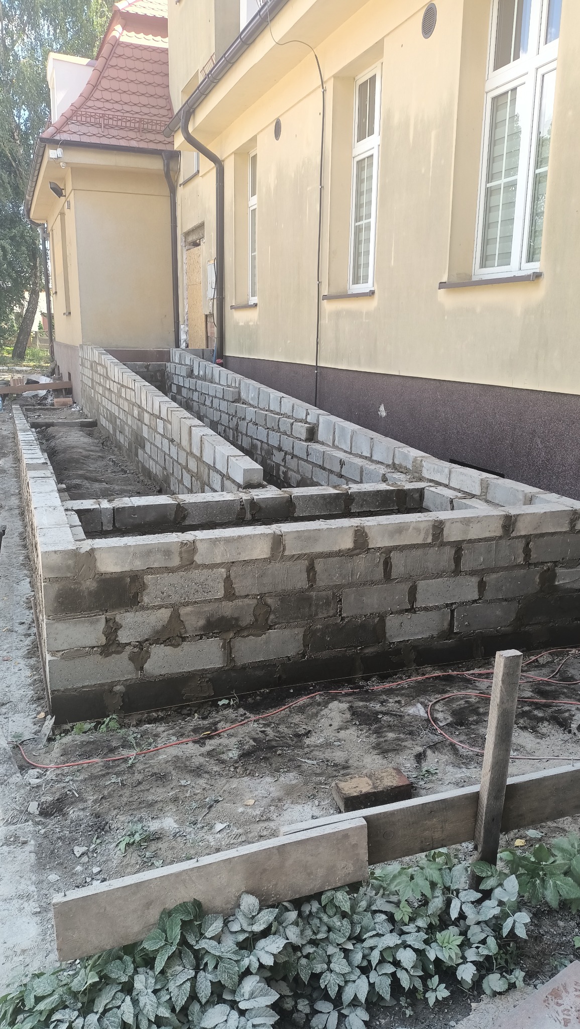 Wznoszenie ścian fundamentowych z bloczków betonowych przy budynku, widoczne zaprawy i roślinność wokół