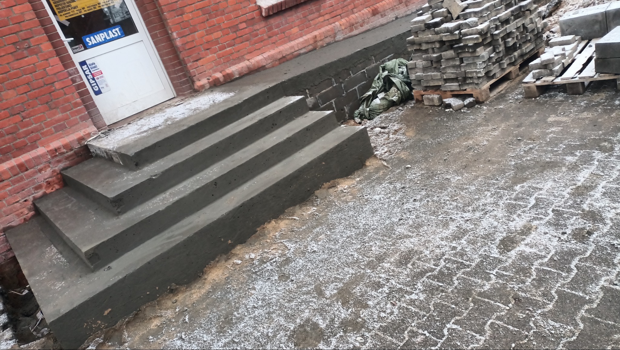 Nowe, surowe betonowe schody wejściowe do budynku z czerwonej cegły, z paletami kostki brukowej obok, w trakcie prac wykończeniowych.