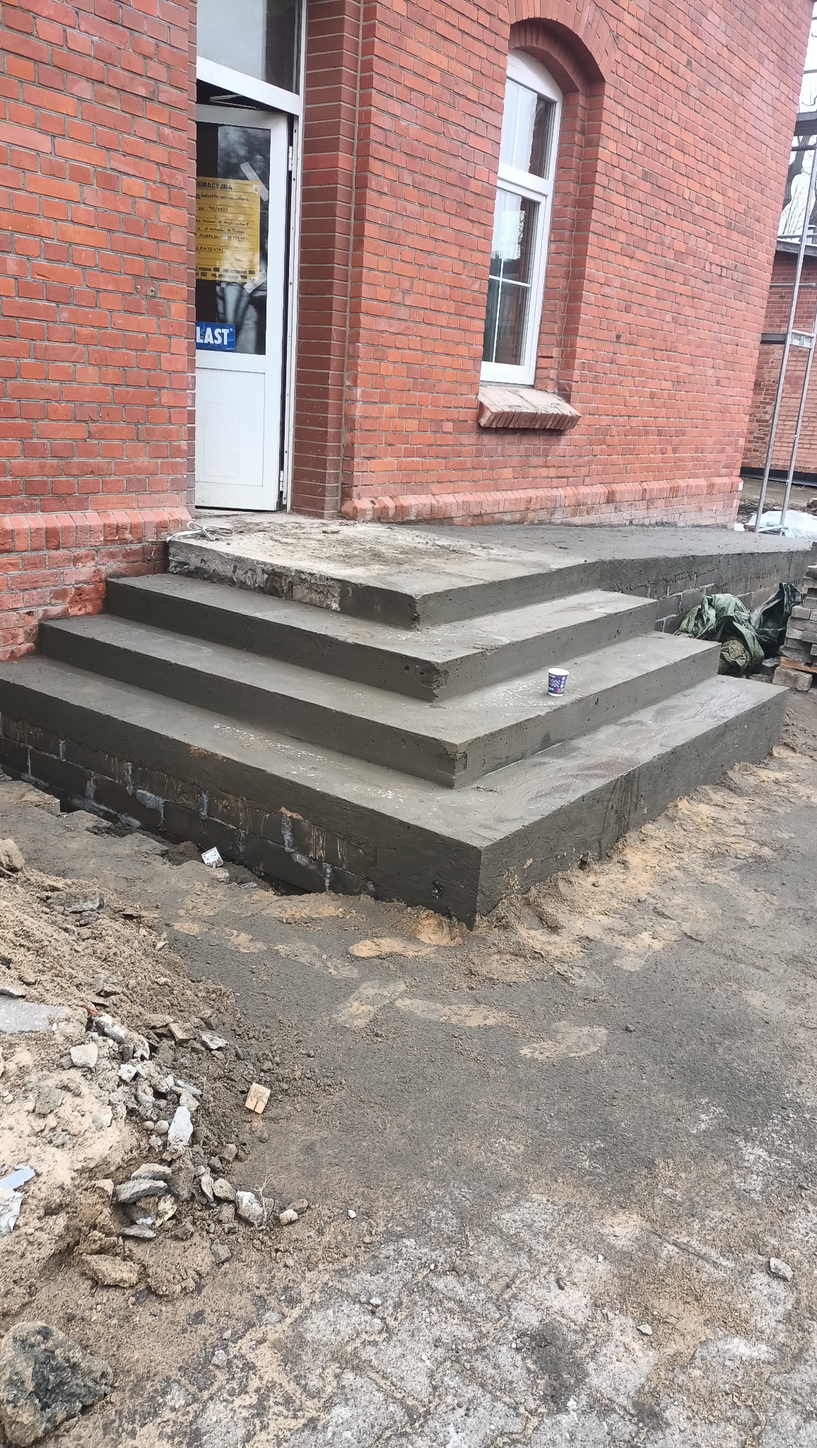 Świeżo wykonane betonowe schody wejściowe do budynku z czerwonej cegły, z widocznym kubkiem po napoju na jednym ze stopni i śladami prac budowlanych wokół.