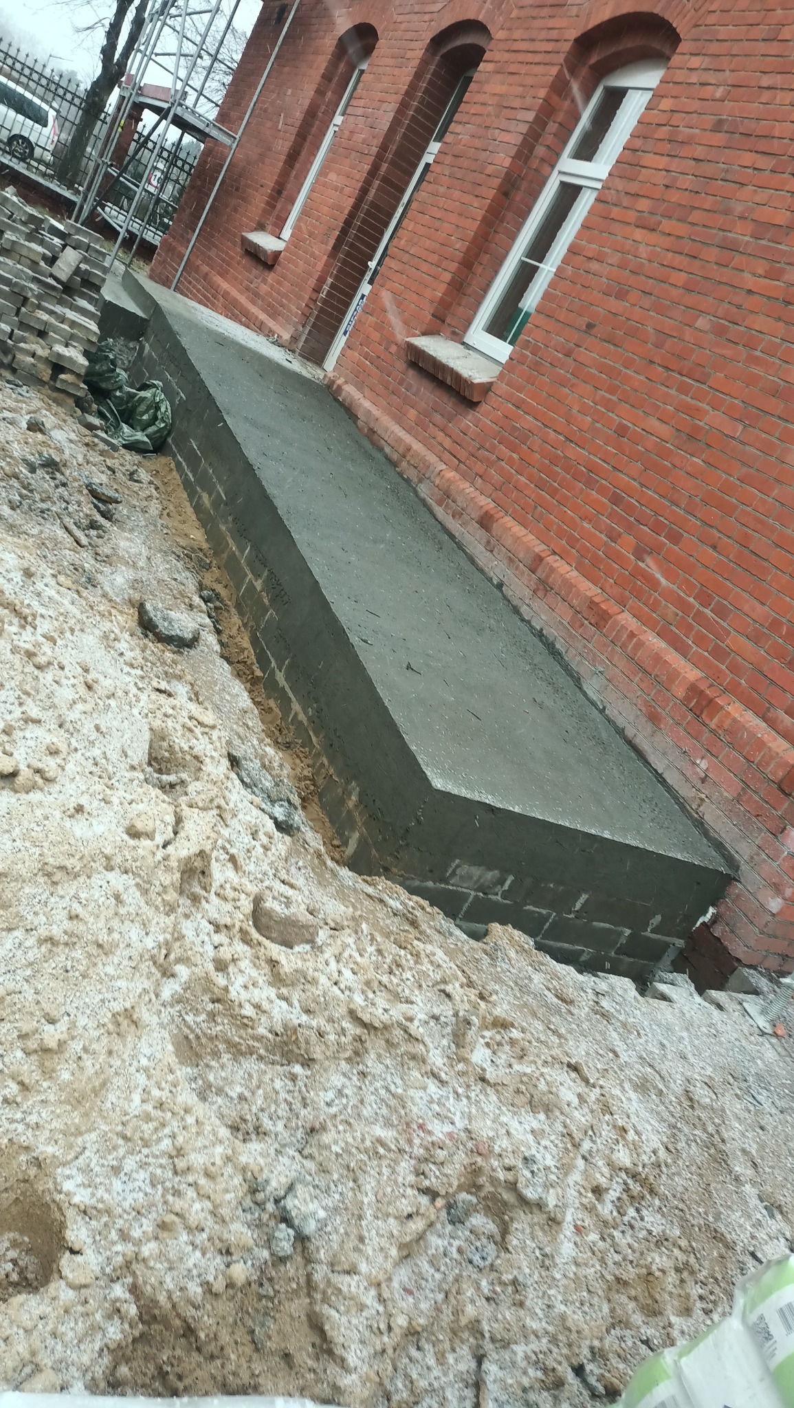 Świeżo wylany betonowy fundament przy ceglanym budynku z białymi oknami, widoczne ślady po szalunku, obok sterta piasku i ułożone cegły, na górze rusztowanie.