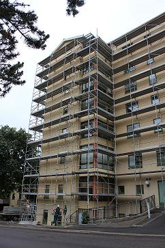 Budynek wielopiętrowy w trakcie remontu, całkowicie otoczony metalowym rusztowaniem, z widocznymi pracownikami na najniższym poziomie.