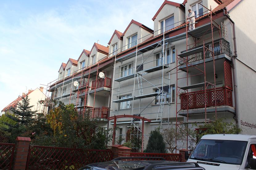 Malowanie elewacji budynku wielorodzinnego z użyciem rusztowań, widoczny pracownik w białym kombinezonie malujący górną część budynku.