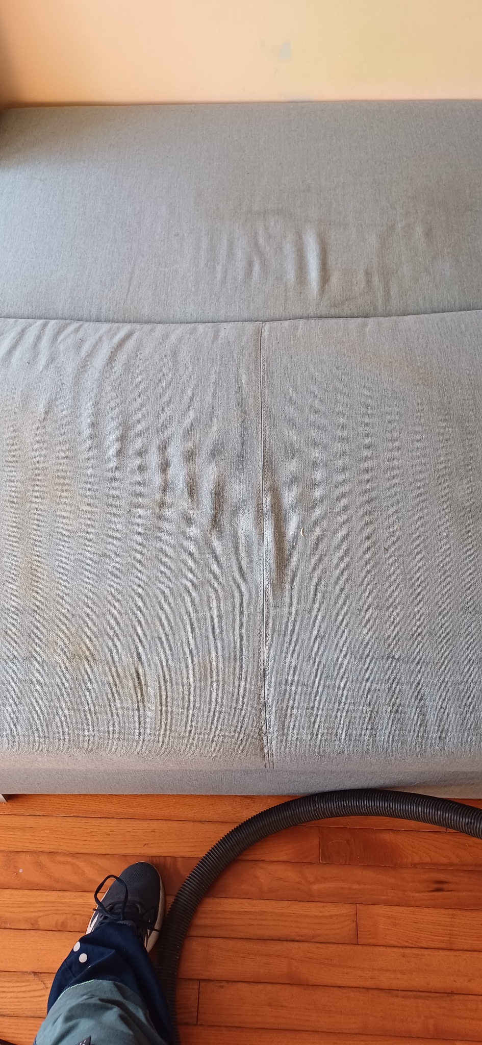 Fragment sofy z widocznymi plamami przed czyszczeniem, rura odkurzacza do prania tapicerki i noga operatora w kadrze.