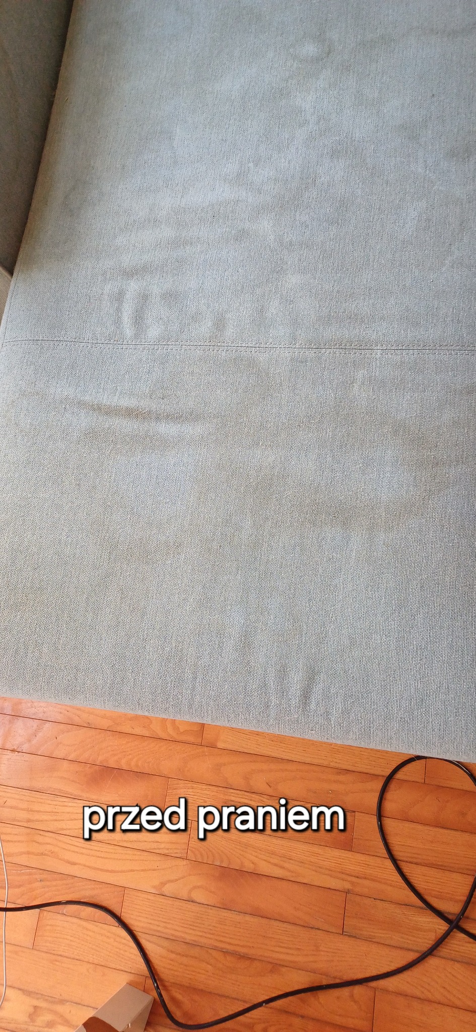 Widok z góry na szarą kanapę z widocznymi plamami i zabrudzeniami, z napisem 'przed praniem' na drewnianej podłodze w dolnej części kadru.