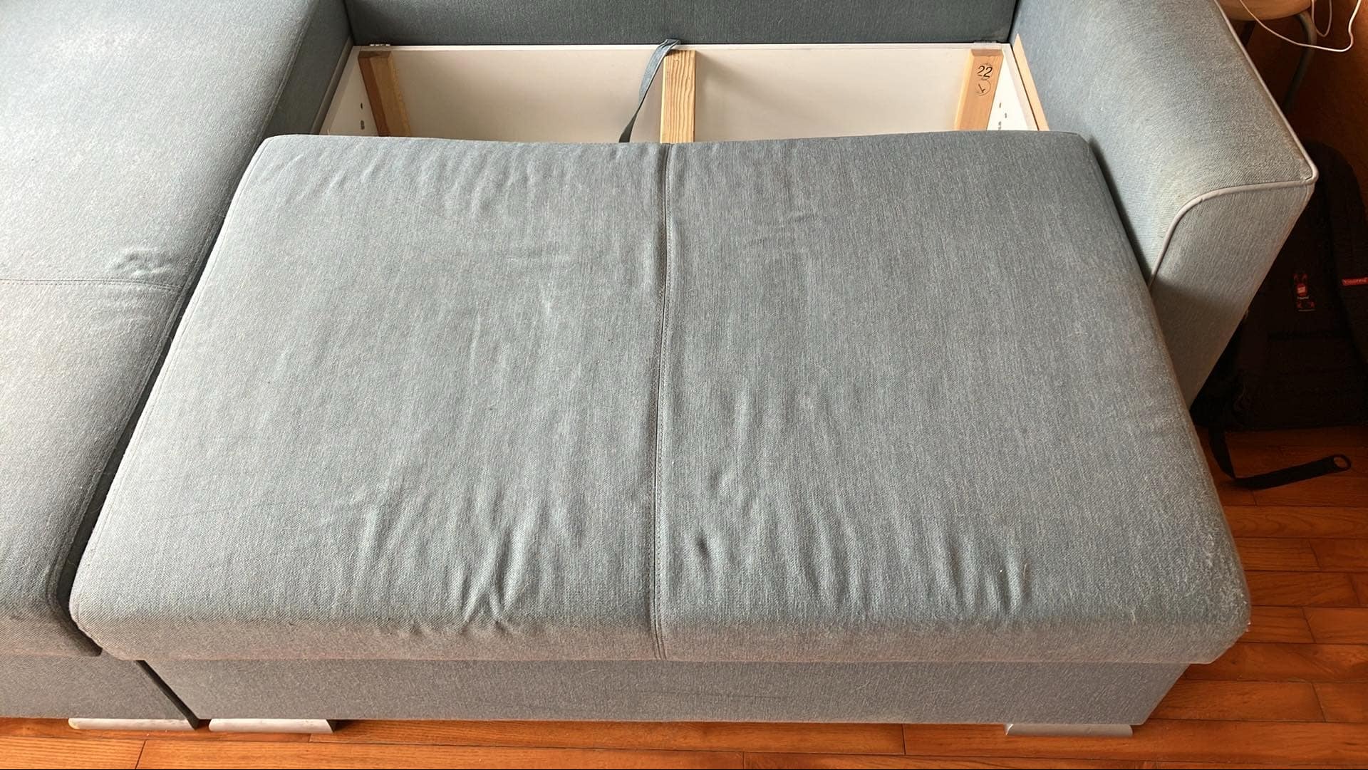 Rozłożona szara kanapa z widocznym pojemnikiem na pościel i drewnianą konstrukcją wewnątrz, na tle drewnianej podłogi.