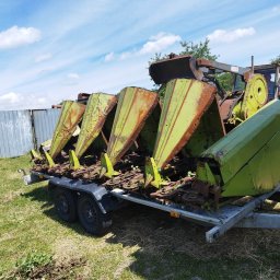Usługi transportowe i handlowe Oleksandr Horyn - Przyczepa z zamontowanym, używanym zespołem żniwnym do kukurydzy w kolorze zielonym i żółtym, z widocznymi śladami rdzy, zaparkowana na trawie w słoneczny dzień.