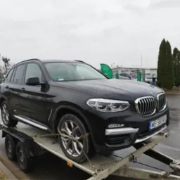 Czarny samochód BMW X3 zabezpieczony pasami transportowymi na metalowej platformie lawety, widok z boku, mokra nawierzchnia.