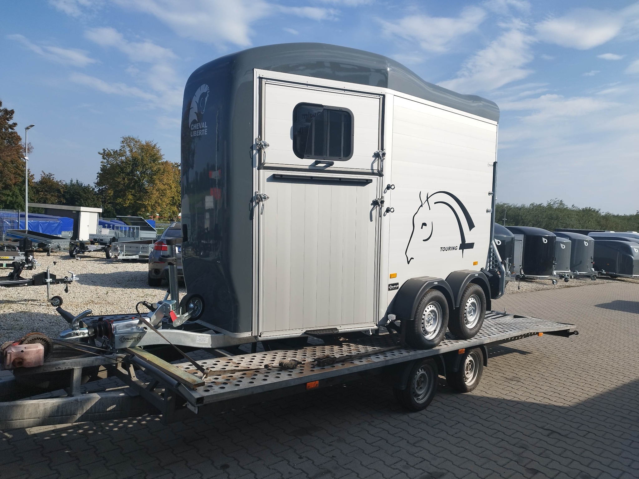Szaro-biała przyczepa do przewozu koni marki Cheval Liberte Touring, umieszczona na platformie lawety, widoczna z boku na tle błękitnego nieba i innych przyczep.