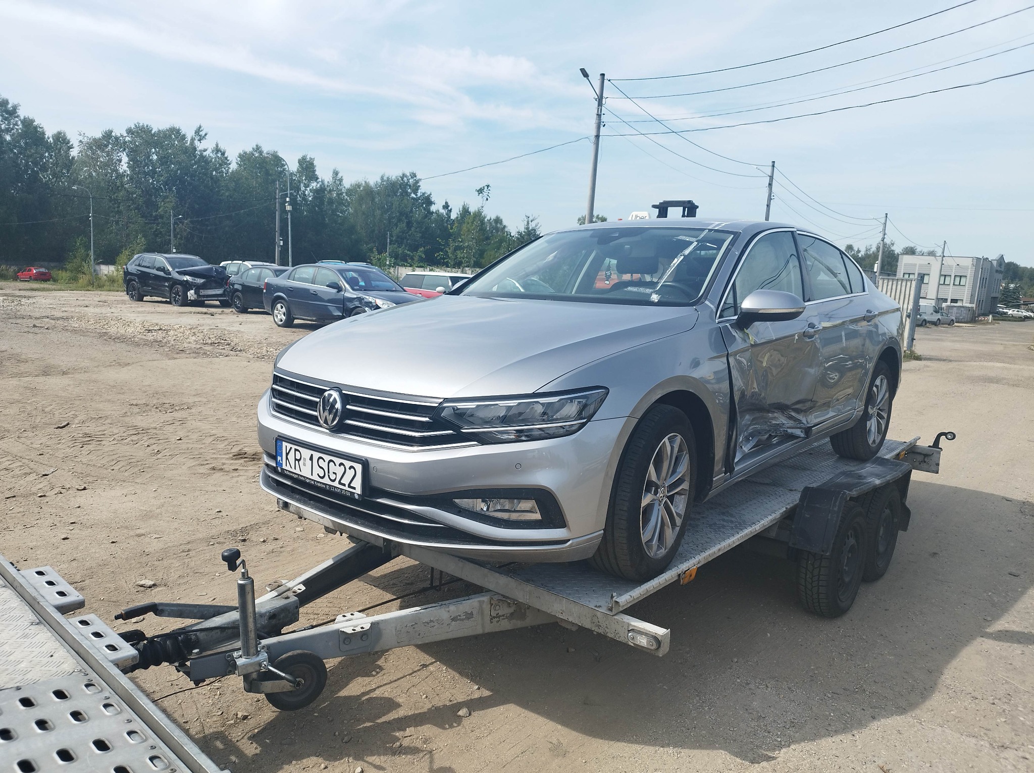 Uszkodzony srebrny Volkswagen Passat na metalowej lawecie z widocznymi uszkodzeniami karoserii po stronie kierowcy, tablica rejestracyjna KR 1SG22, na szutrowym parkingu z innymi pojazdami w tle.