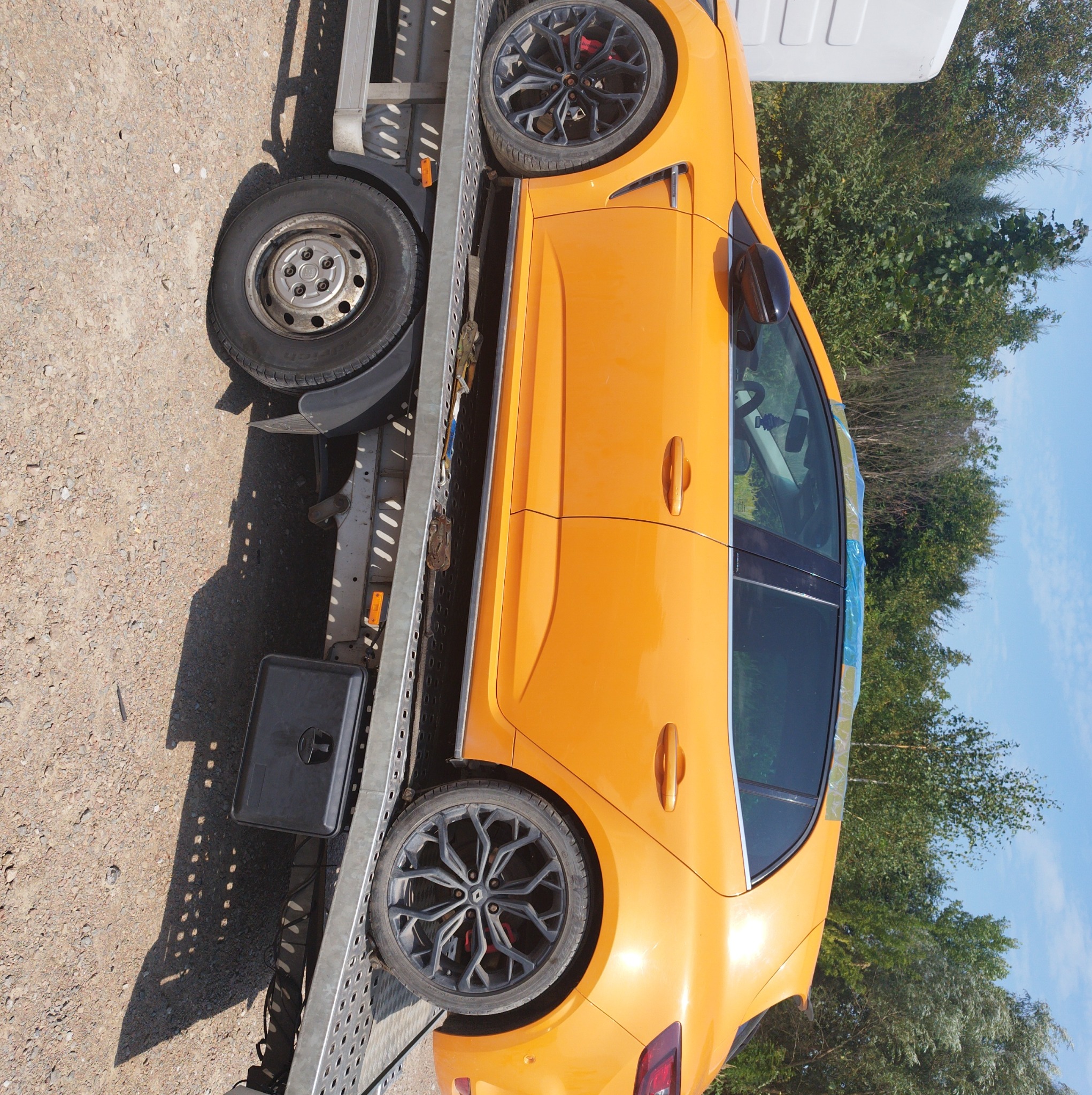 Pomarańczowy samochód sportowy, zabezpieczony folią, na metalowej platformie lawety, widok z boku, teren żwirowy i drzewa w tle.