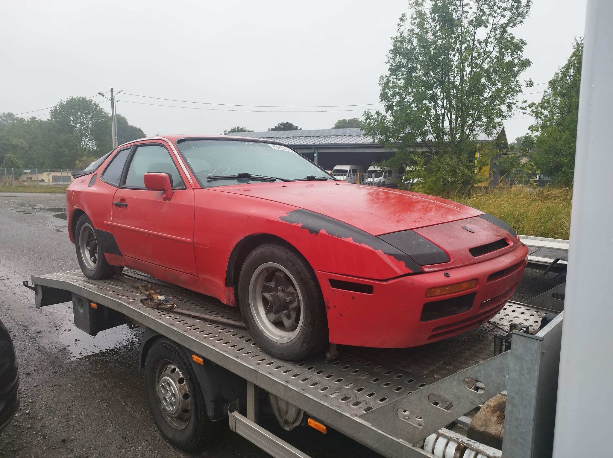 Czerwone Porsche 944 na lawecie z widocznymi śladami użytkowania i odchodzącą farbą, transportowane w pochmurny dzień.