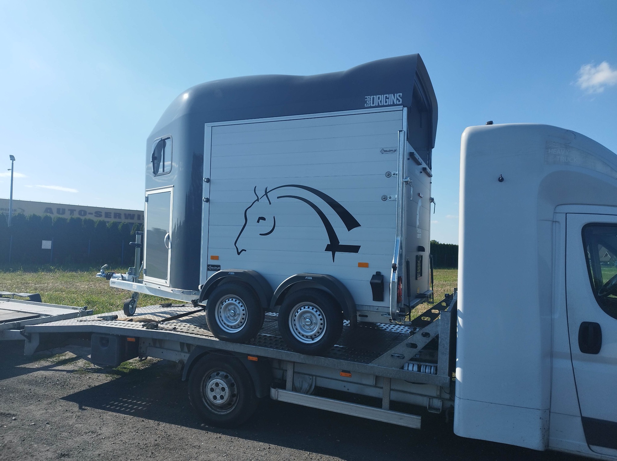 Przyczepa do przewozu koni marki Origins, umieszczona na platformie lawety, widoczna z boku, z grafiką konia na białej części bocznej.