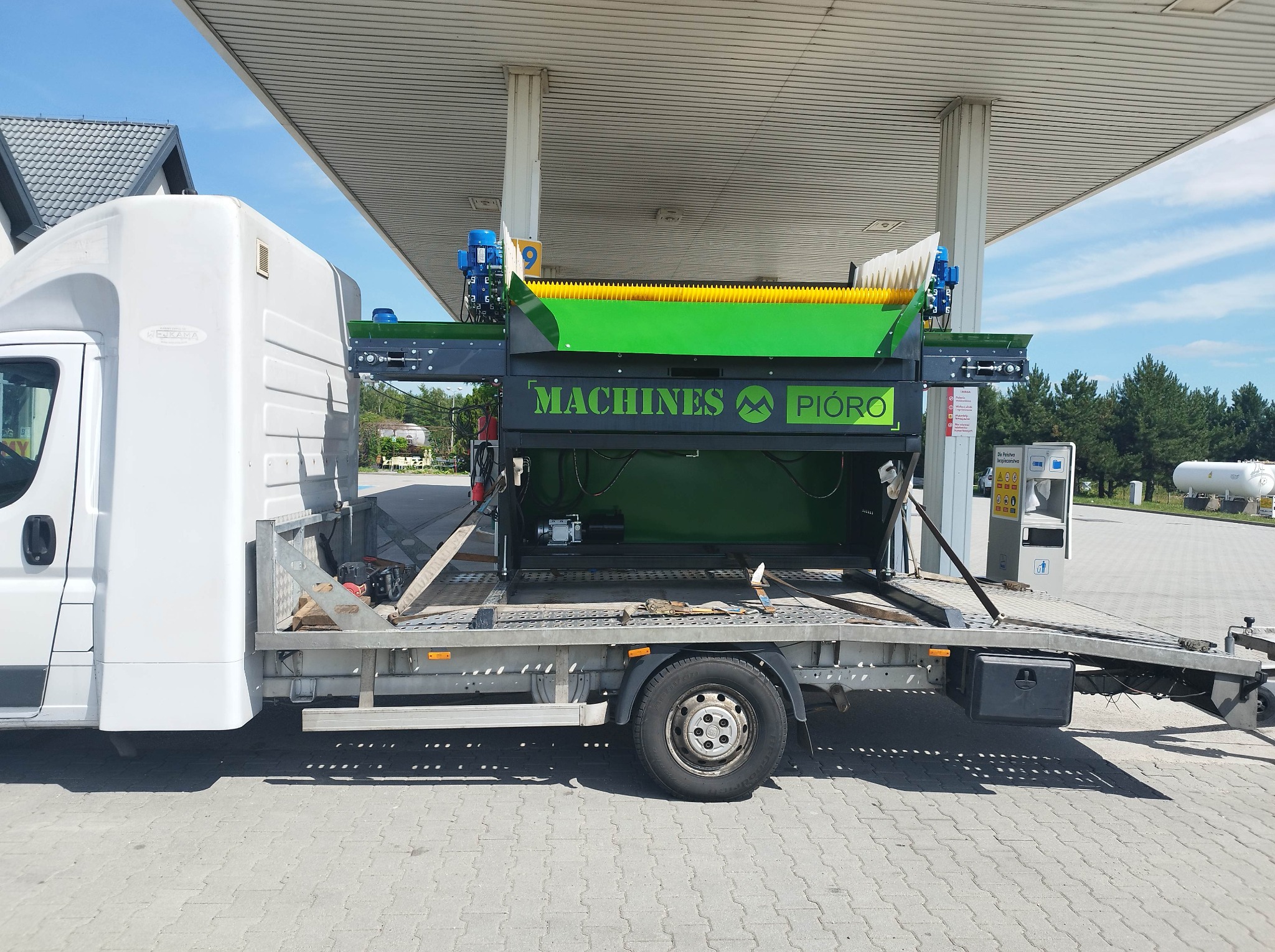 Biały samochód dostawczy z platformą transportową przewożący zielono-czarną maszynę z logo 'Machines Pióro' na tle stacji benzynowej.