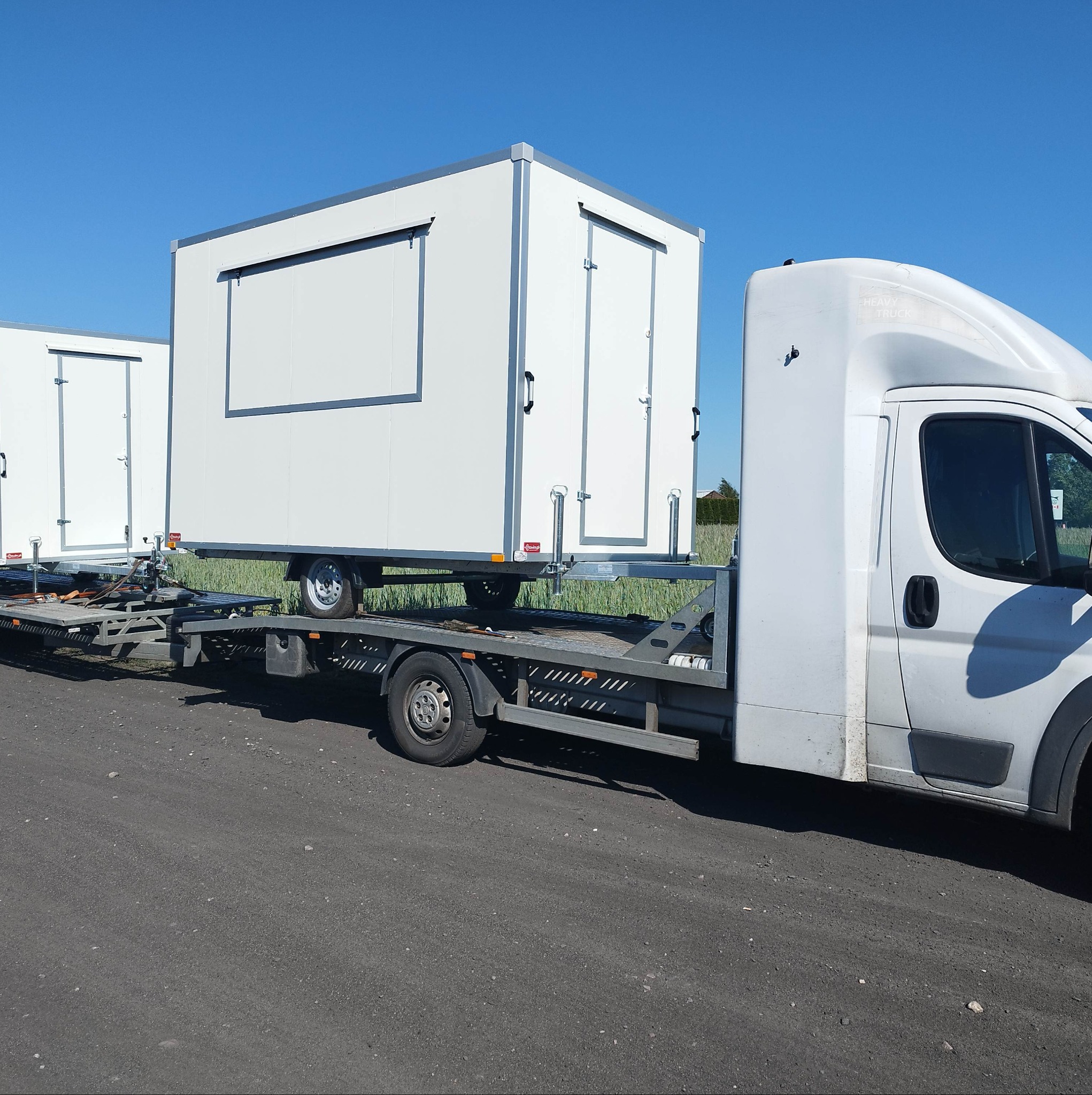 Biały bus z platformą transportową przewozi dwa białe kontenery z drzwiami, na tle błękitnego nieba.