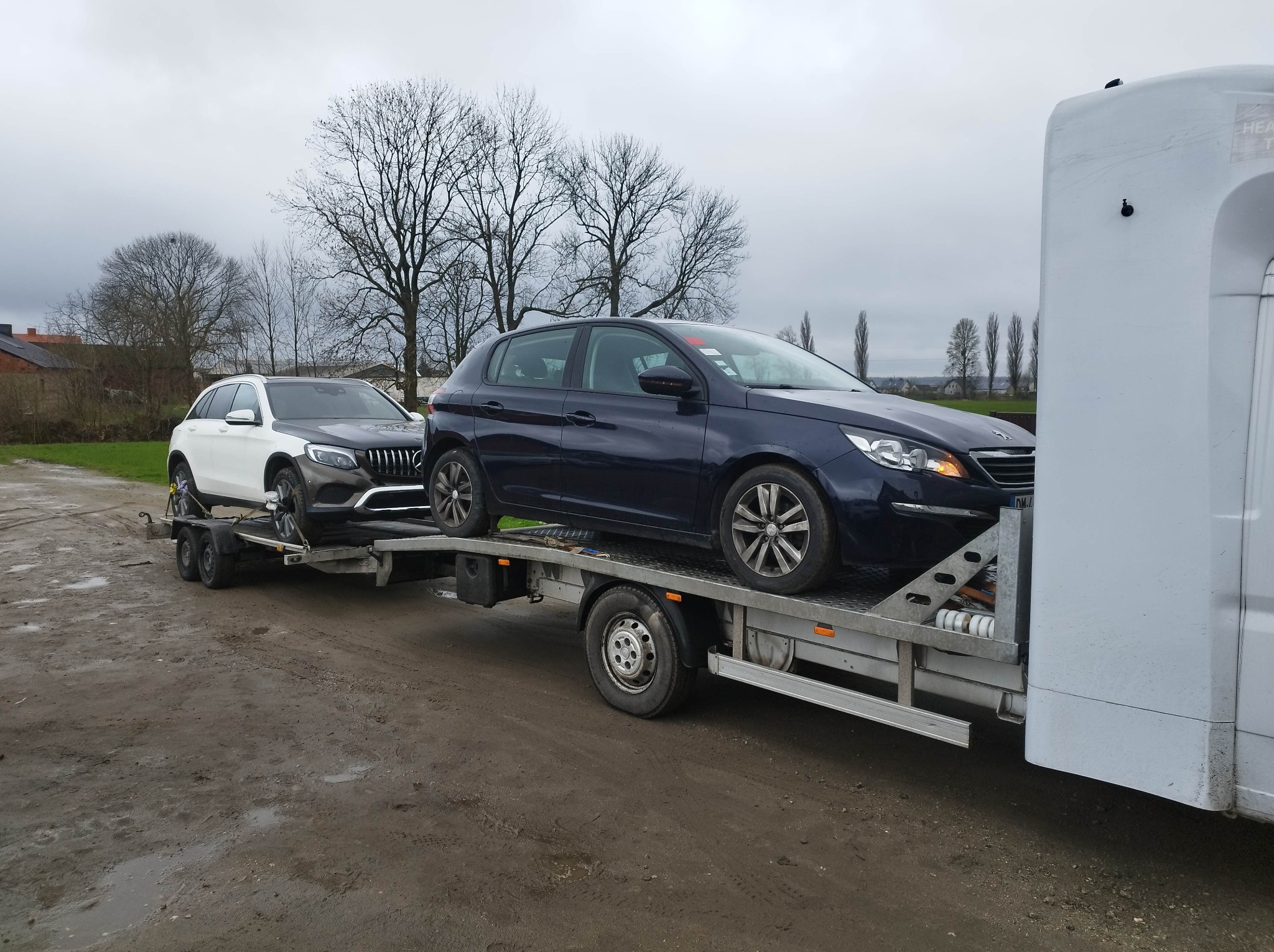 Dwa samochody osobowe, biały Mercedes i granatowy Peugeot, załadowane na lawetę w pochmurny dzień. Mercedes na małej przyczepie doczepionej do głównej lawety.