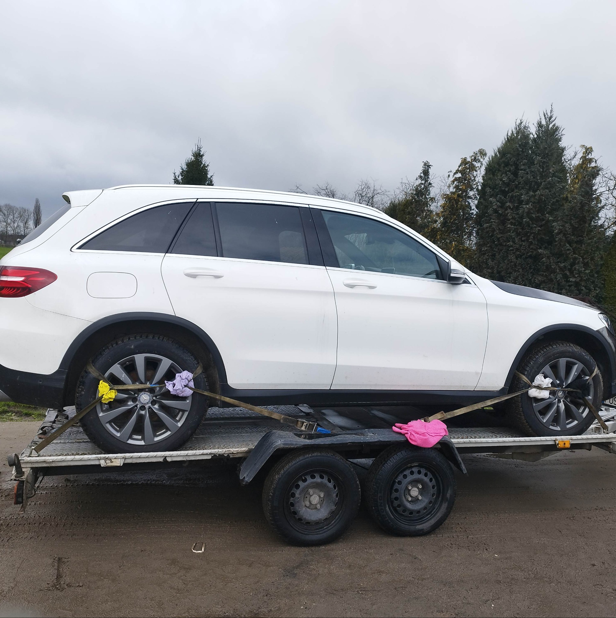 Biały samochód osobowy Mercedes-Benz zabezpieczony pasami transportowymi, umieszczony na przyczepie lawety, widok z boku na tle pochmurnego nieba i drzew.