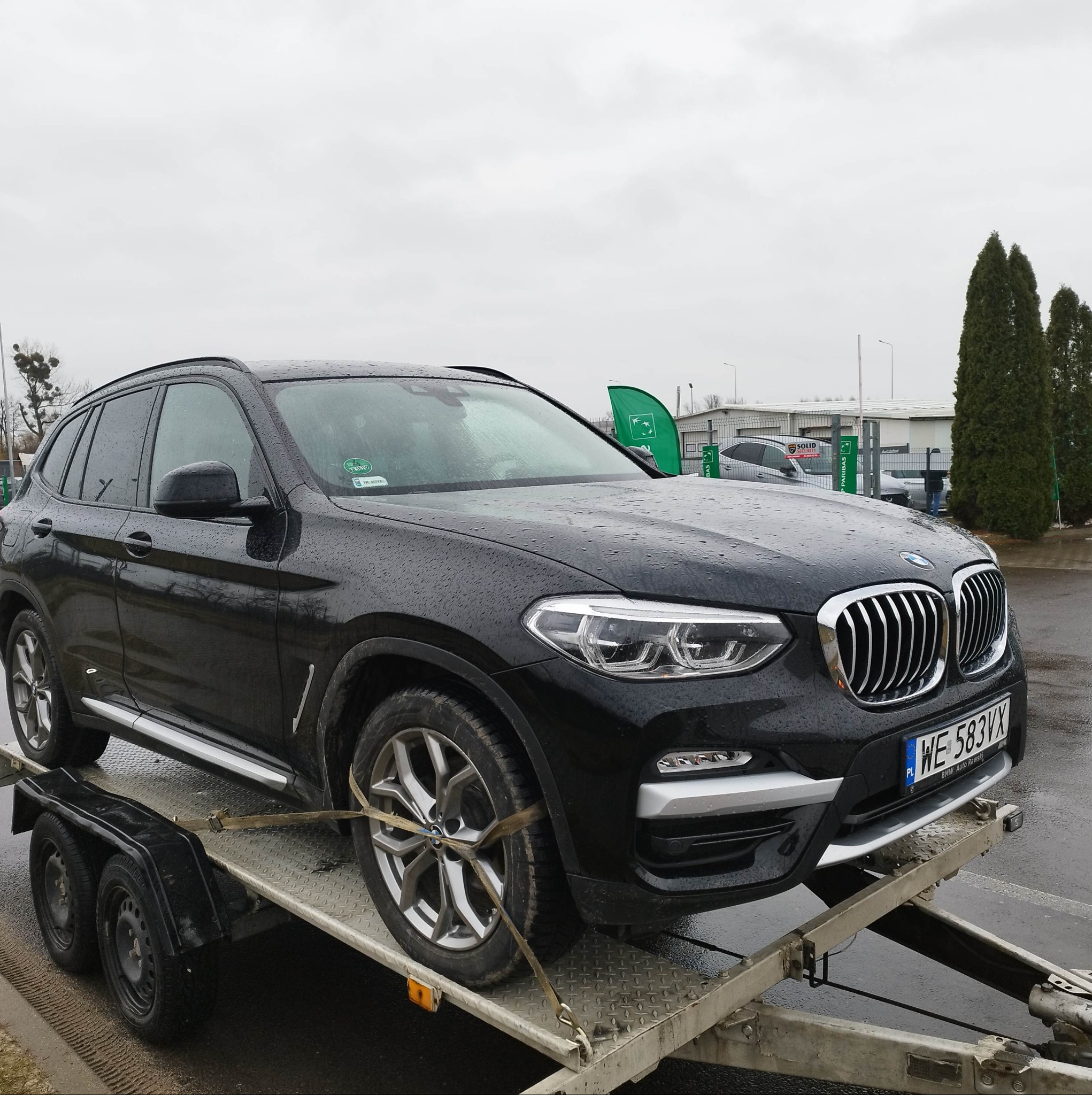 Czarny samochód BMW X3 zabezpieczony pasami transportowymi na metalowej platformie lawety, widok z boku, mokra nawierzchnia.