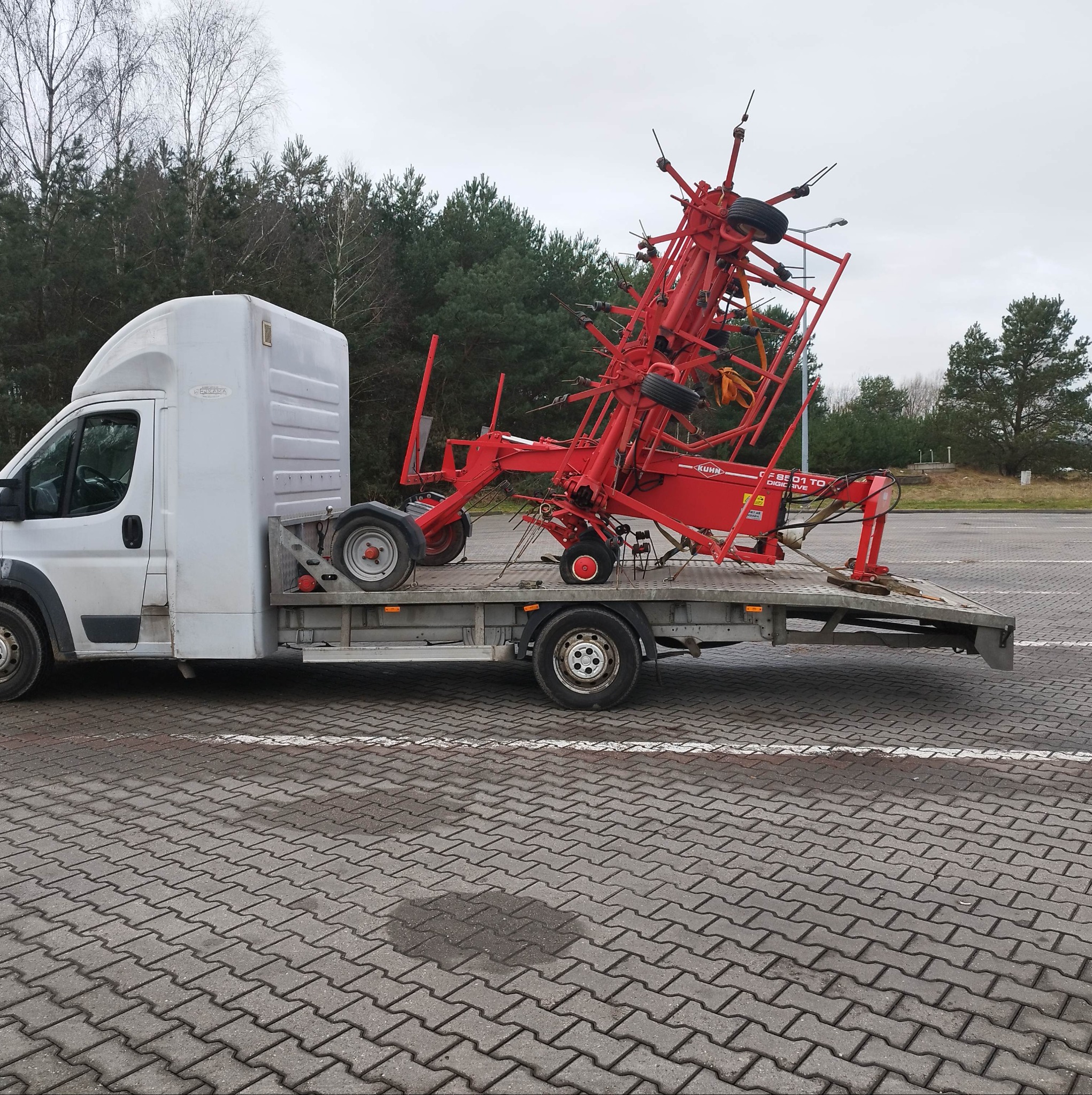 Biały samochód dostawczy z platformą transportową przewożący czerwoną przetrząsarkę karuzelową Kuhn na parkingu z kostki brukowej.