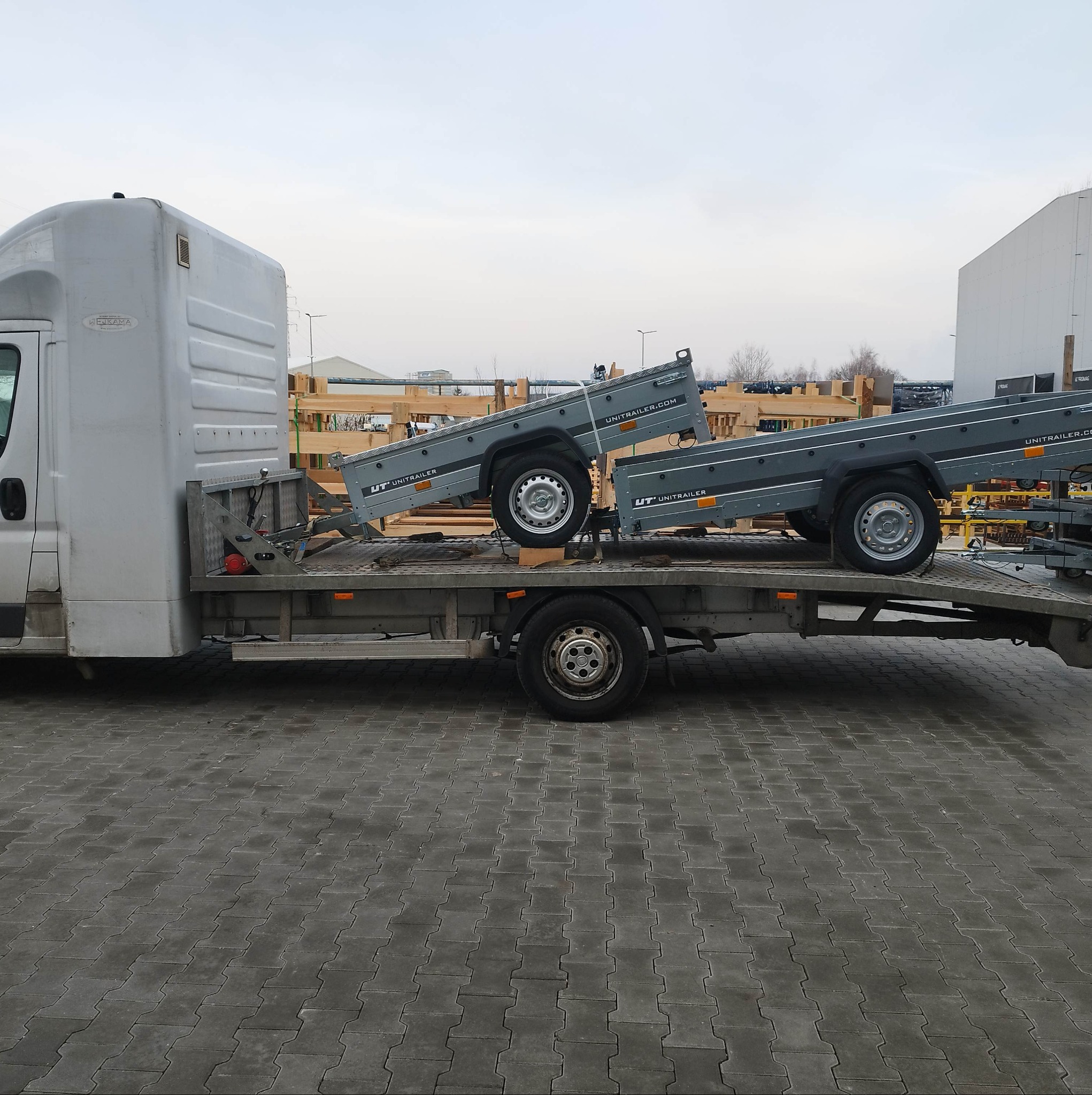 Srebrny samochód dostawczy z platformą transportową przewozi dwie nowe, szare przyczepy Unitrailer na tle szarej kostki brukowej i pochmurnego nieba.