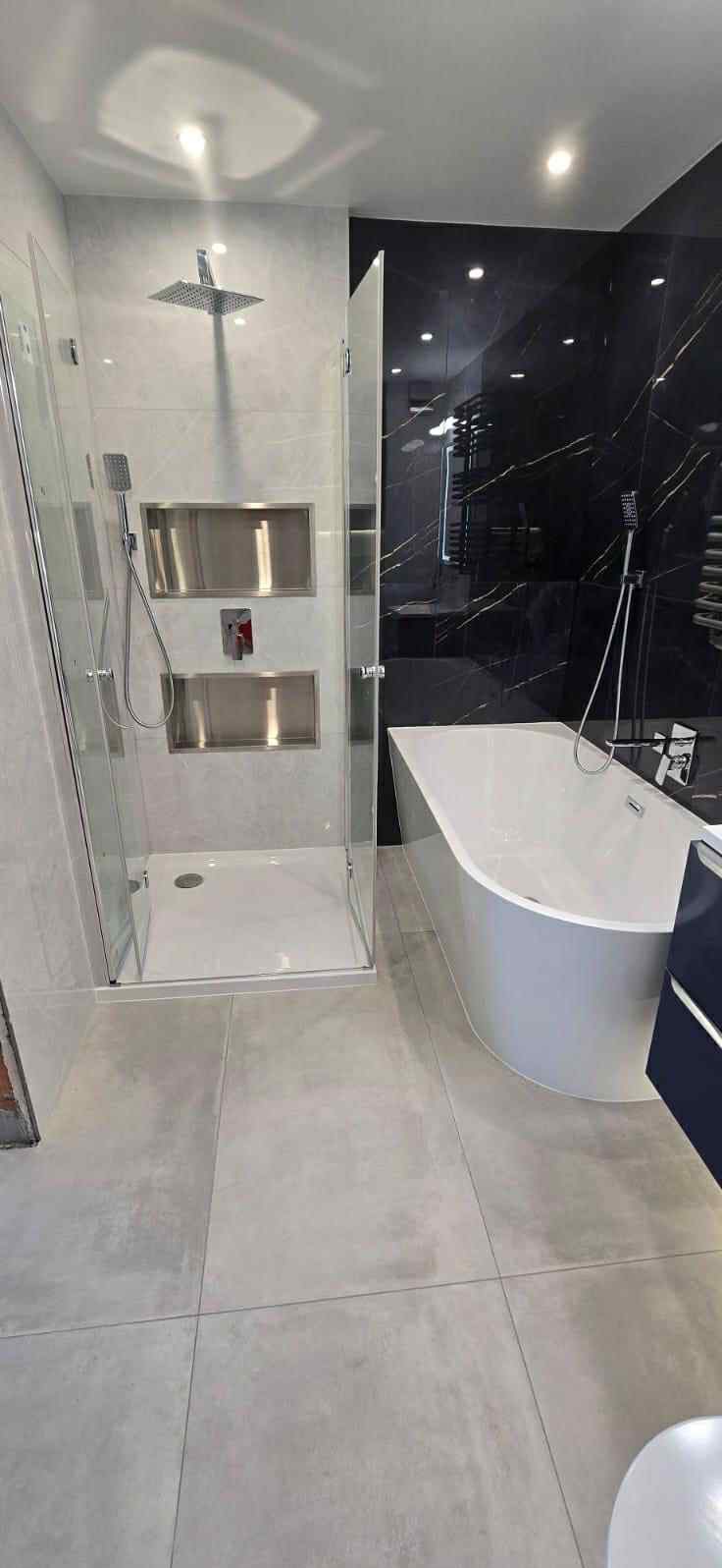 Nowoczesna łazienka po remoncie: kabina prysznicowa ze srebrnymi wnękami, wanna przy czarnej ścianie z marmurkowym wzorem, szare płytki podłogowe.