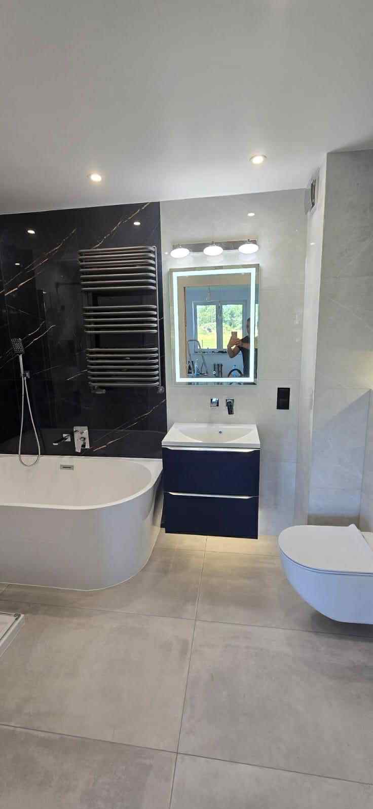 Nowoczesna łazienka z wanną, umywalką w szafce, podświetlanym lustrem i grzejnikiem drabinkowym na tle czarnej ściany z marmurowym wzorem.