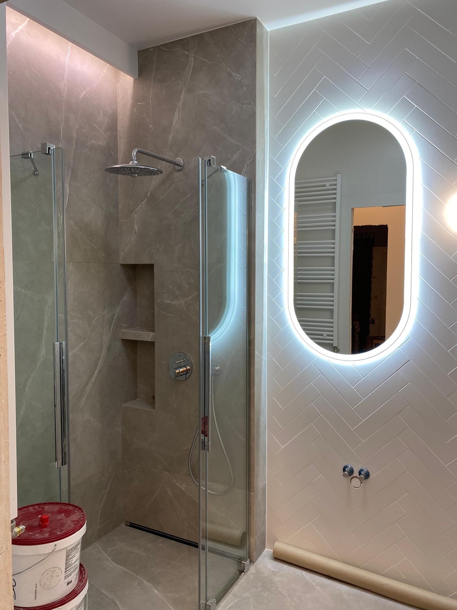 Wykończona łazienka: kabina prysznicowa z szarymi płytkami imitującymi marmur, ściana z białymi płytkami ułożonymi w jodełkę i podświetlane lustro owalne.