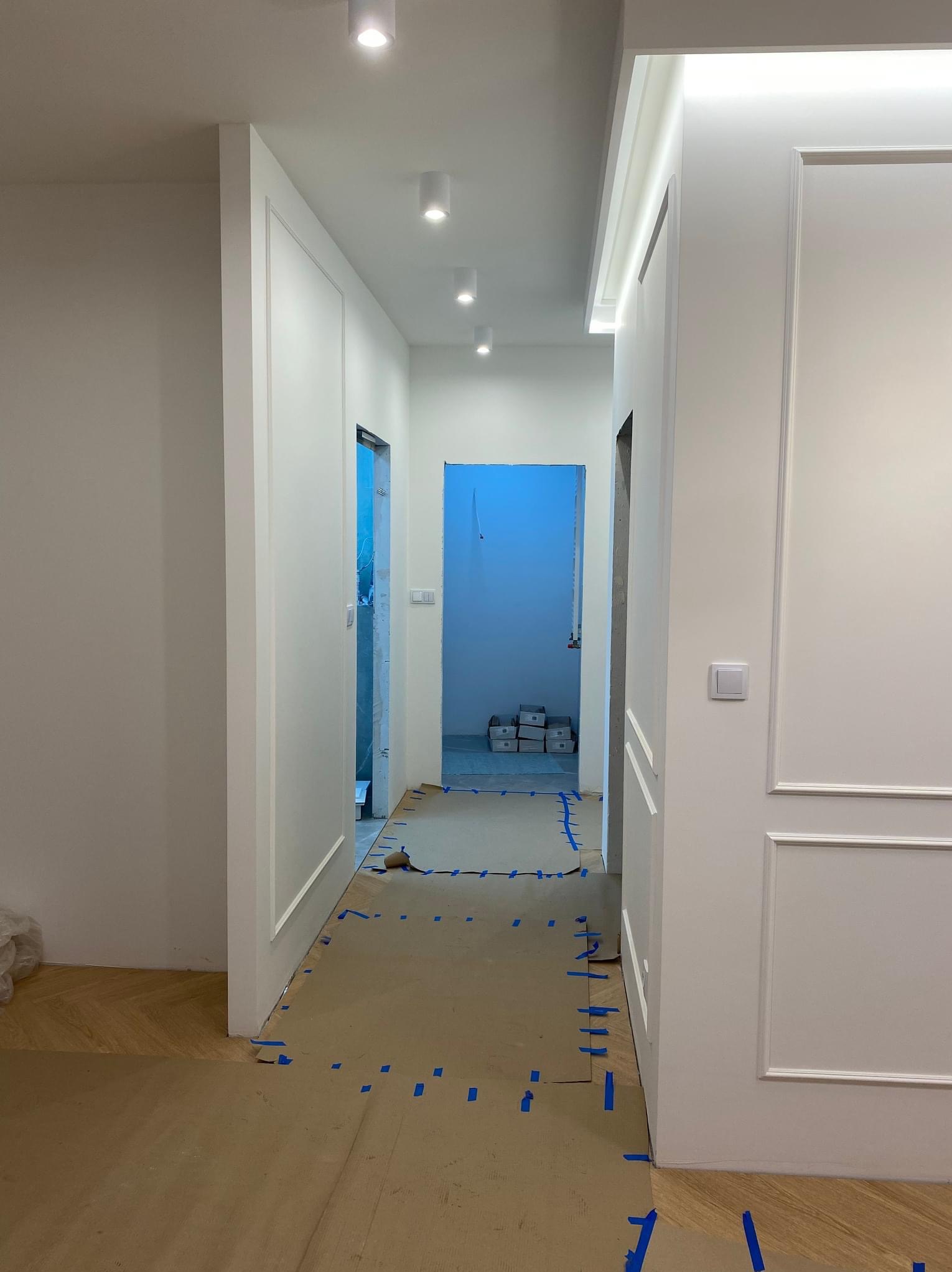 Korytarz w trakcie prac wykończeniowych, zabezpieczone podłogi, widoczne listwy dekoracyjne i oświetlenie sufitowe.