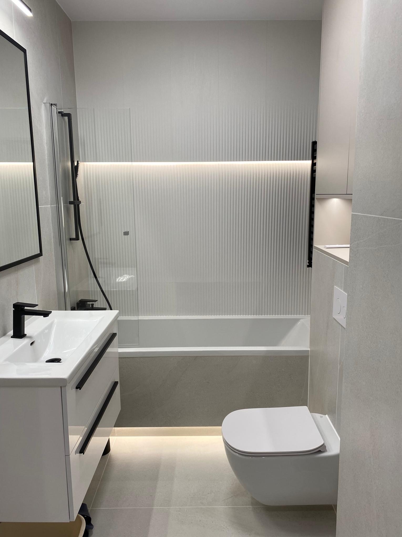 Nowoczesna łazienka w odcieniach szarości z podświetleniem LED pod wanną i nad nią. Umywalka z czarną armaturą i wisząca toaleta.