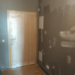 Pokój w trakcie szpachlowania i malowania ścian na szaro, zabezpieczone folią drzwi i podłoga, widoczne ślady po szpachlowaniu.
