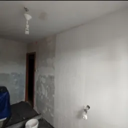 Pokój w trakcie remontu: ściana częściowo wyszpachlowana, fragment pomalowany na biało z fakturą, widoczne zabezpieczenia i narzędzia malarskie. Przygotowanie do malowania.