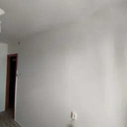 Jasne, świeżo pomalowane ściany w pokoju z widocznym gniazdkiem i lampą sufitową. Widoczny fragment podłogi i drzwi. Minimalistyczne wnętrze po remoncie.