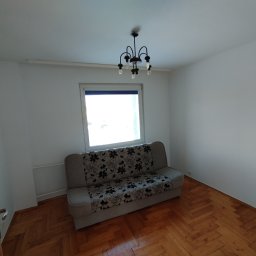 Mateusz Rzepa - Pusty pokój z parkietem i sofą w kwiaty pod oknem, po malowaniu ścian na biało. Widoczny żyrandol i fragment drewnianych drzwi.