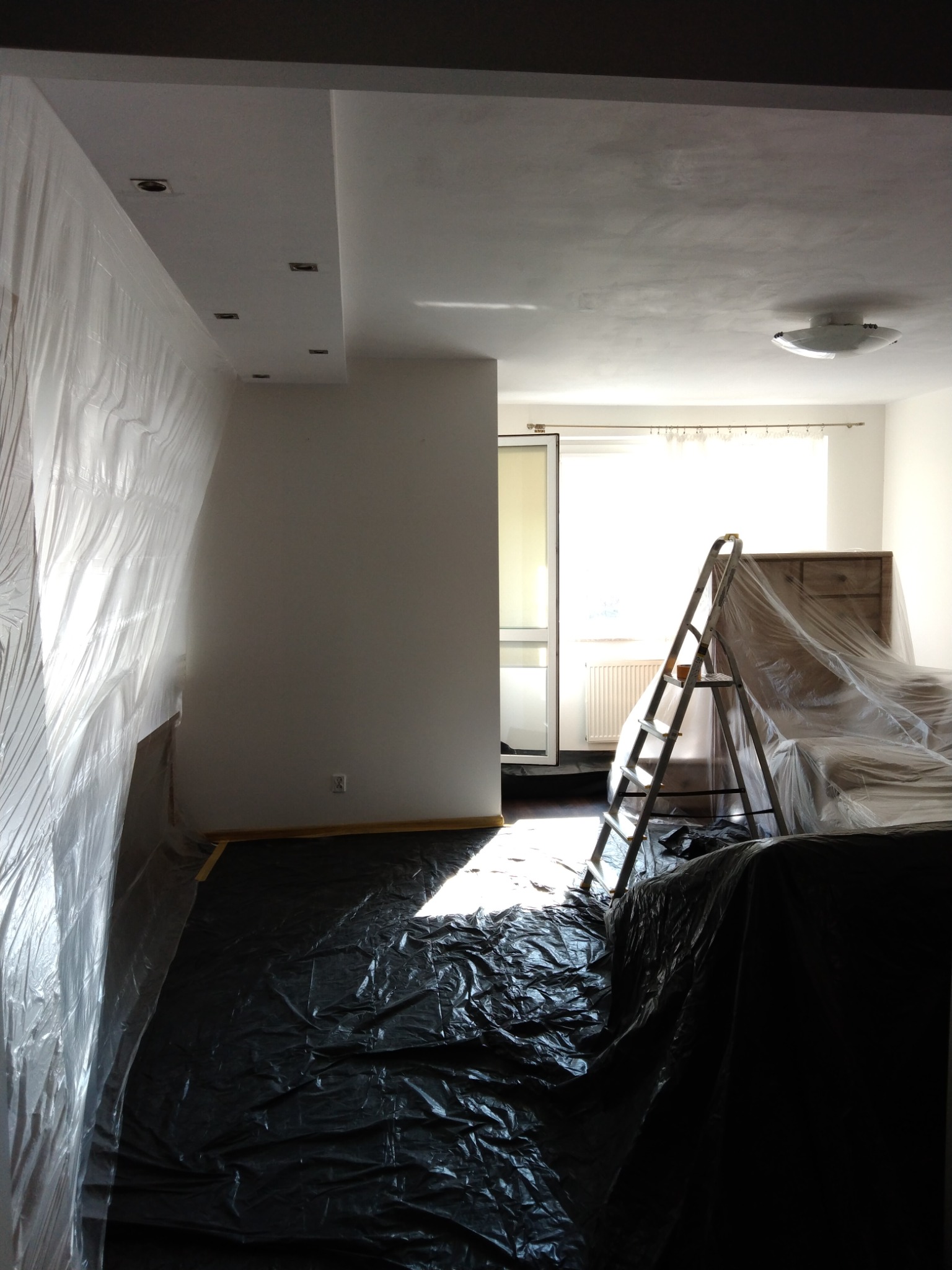 Pokój przygotowany do remontu: ściany i meble zabezpieczone folią malarską, na podłodze czarna plandeka, w tle drabina oparta o meble, jasne światło wpadające przez okno balkonowe.