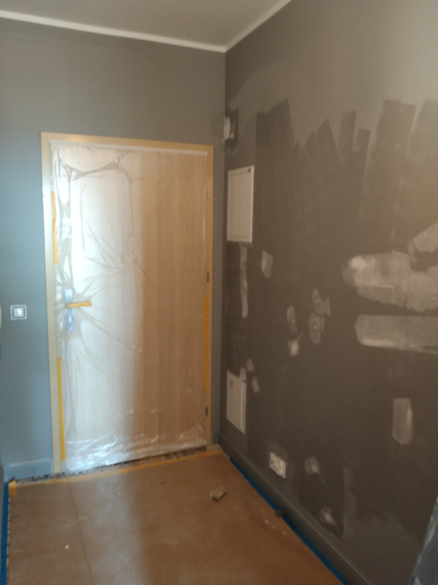 Pokój w trakcie szpachlowania i malowania ścian na szaro, zabezpieczone folią drzwi i podłoga, widoczne ślady po szpachlowaniu.