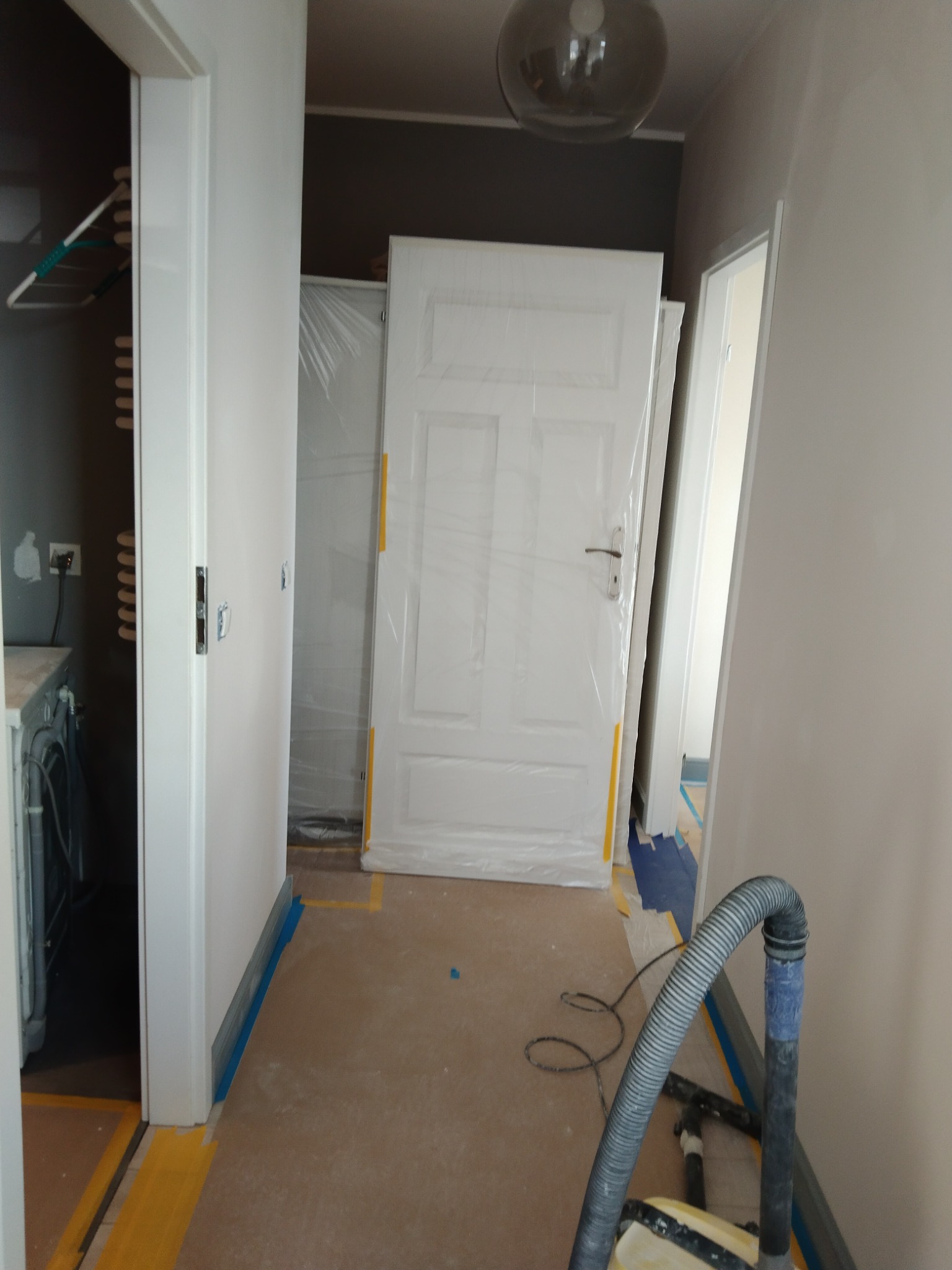Przygotowanie do prac wykończeniowych: zabezpieczone folią drzwi wewnętrzne, podłoga zaklejona taśmą malarską, widoczny odkurzacz przemysłowy i ściany w kolorze jasnego beżu.