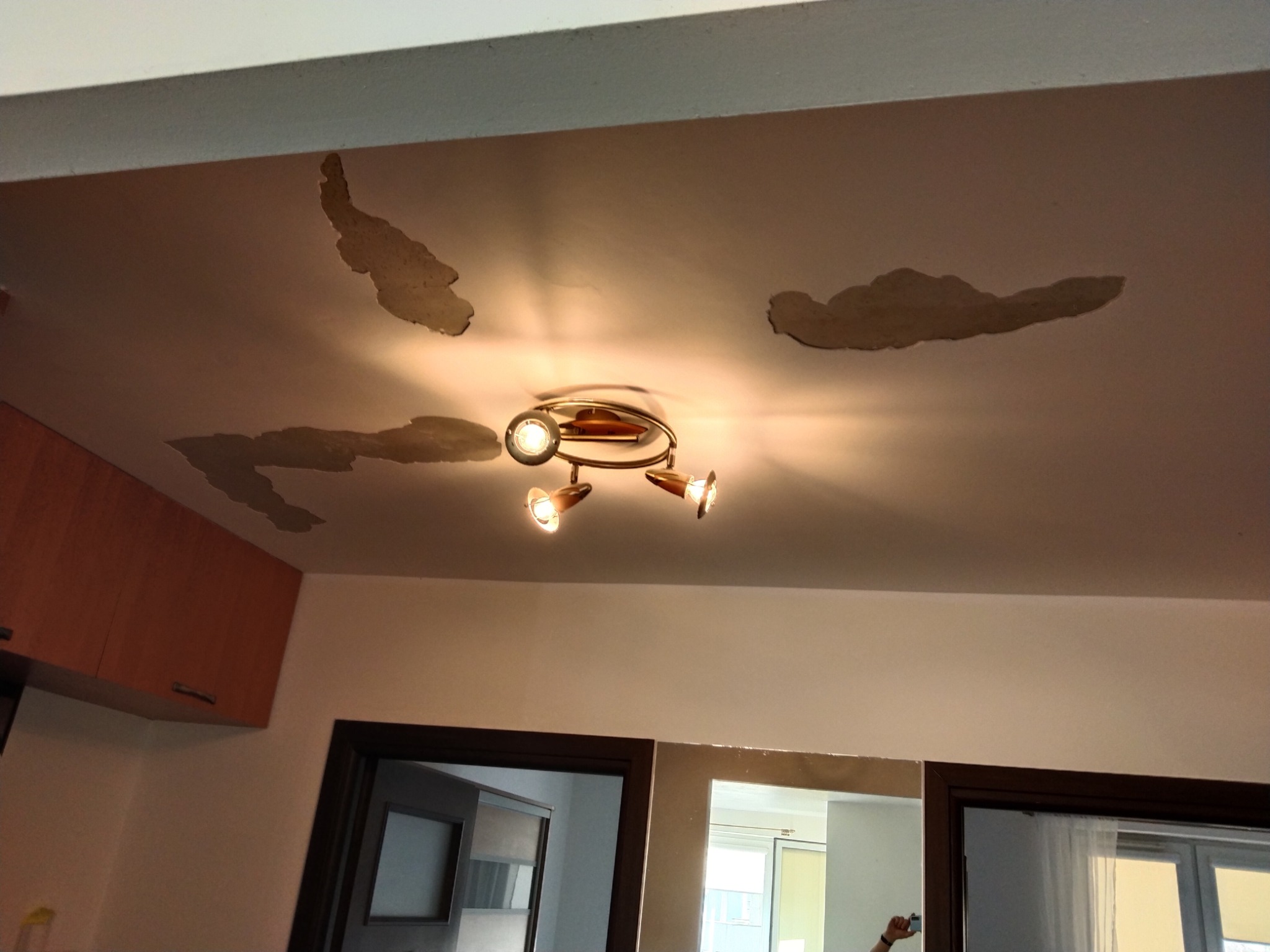 Niedoskonałości na suficie w postaci ubytków tynku o nieregularnych kształtach, oświetlone przez lampę sufitową z trzema reflektorami, widoczne fragmenty pomieszczeń przez otwarte drzwi.