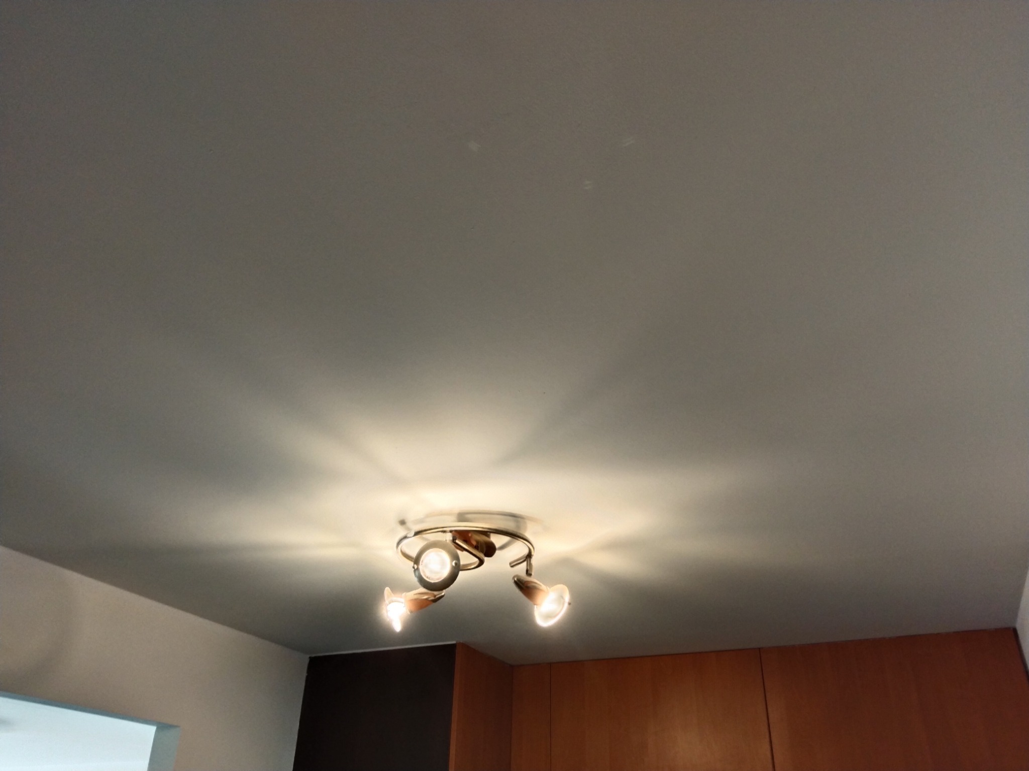 Gładki, pomalowany na szaro sufit z zamontowaną, nowoczesną lampą sufitową z trzema reflektorami, oświetlającymi przestrzeń.