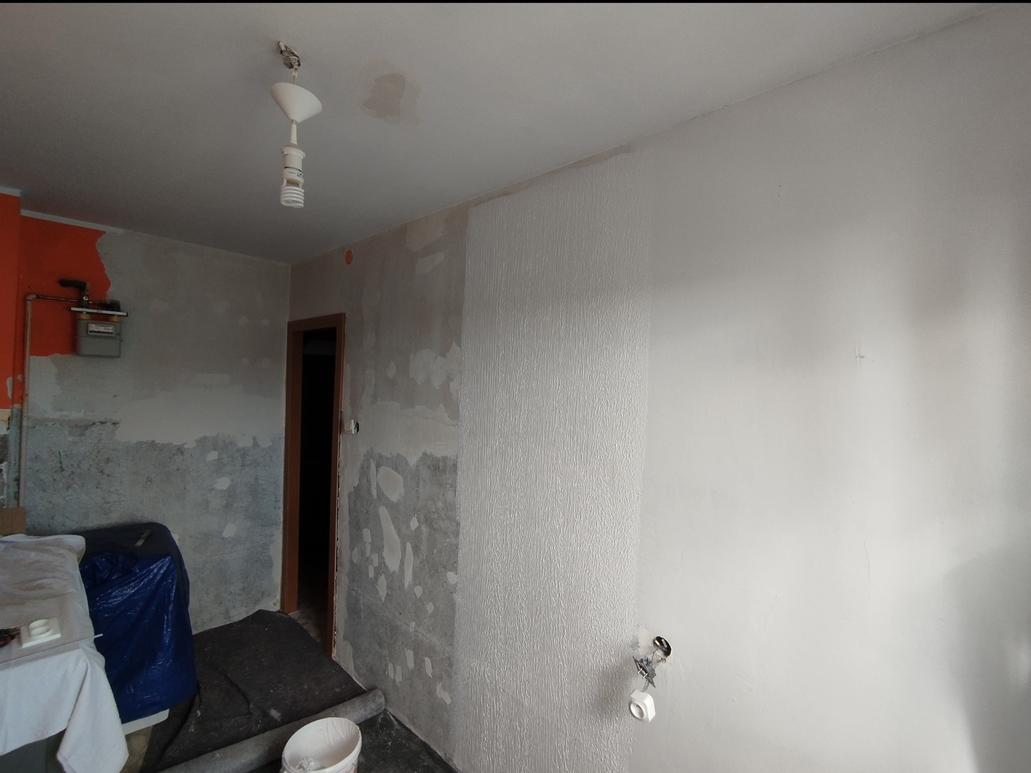 Pokój w trakcie remontu: ściana częściowo wyszpachlowana, fragment pomalowany na biało z fakturą, widoczne zabezpieczenia i narzędzia malarskie. Przygotowanie do malowania.