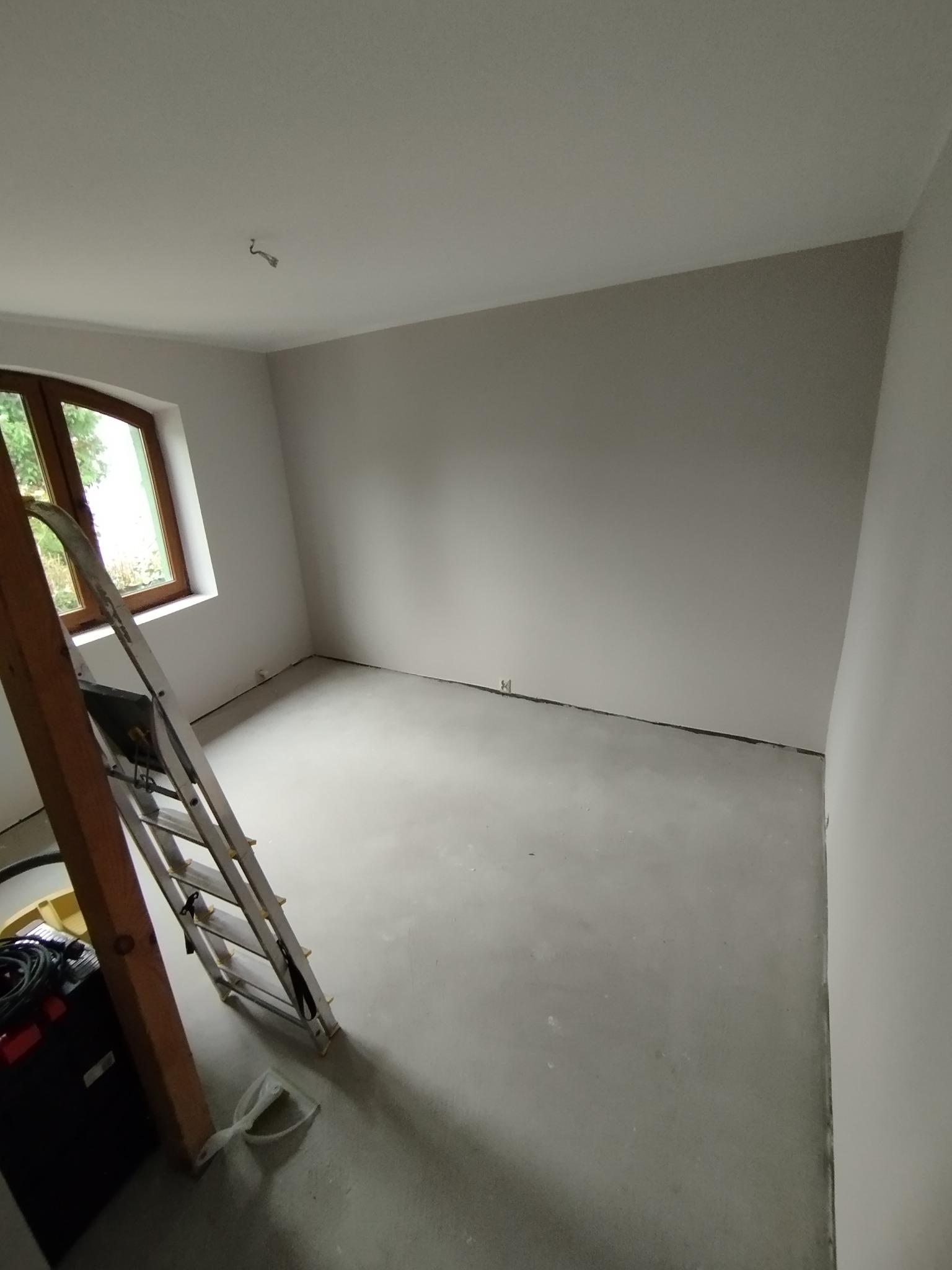 Wnętrze pokoju po szpachlowaniu, przygotowane do malowania. Widoczna drabina, gładkie ściany i sufit, okno z widokiem na zieleń. Minimalistyczny wystrój, surowy stan wykończenia.