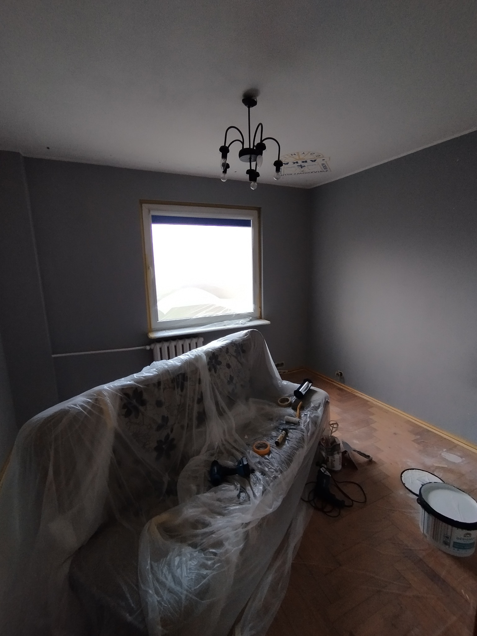 Pokój w trakcie malowania na szaro. Meble zabezpieczone folią, widoczne narzędzia malarskie i puszka z farbą. Przygotowania do odświeżenia wnętrza.