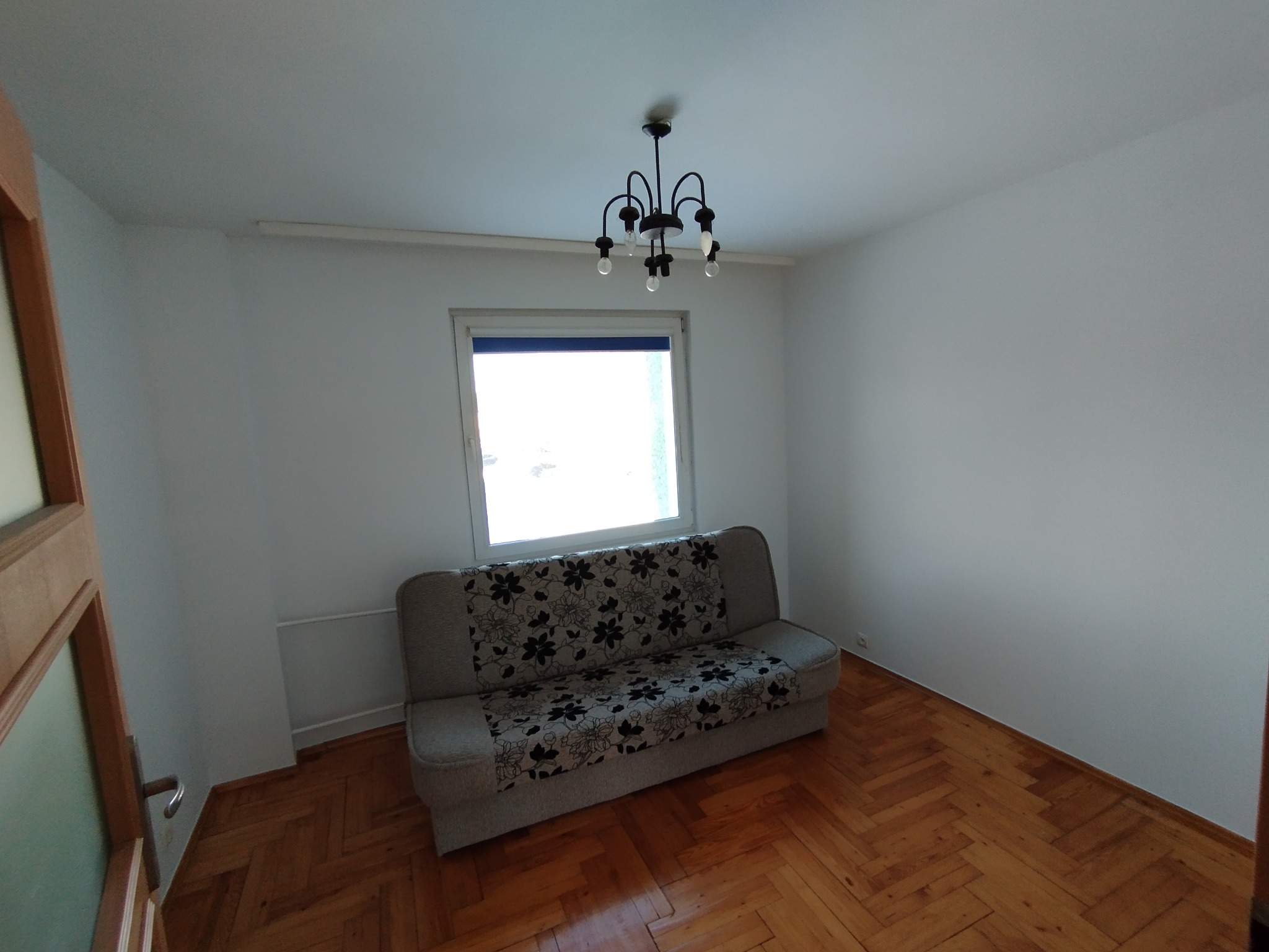 Pusty pokój z parkietem i sofą w kwiaty pod oknem, po malowaniu ścian na biało. Widoczny żyrandol i fragment drewnianych drzwi.